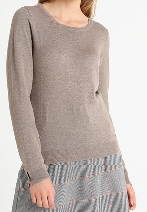 Kobieta ubrana w taupe'owy sweter z okrągłym dekoltem i długimi rękawami, zestawiony z szarą spódnicą w kratę z cienkimi czerwonymi liniami.
