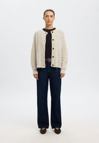 Jasnobeżowy dziergany kardigan z dużymi brązowymi guzikami, noszony na czarnej bluzce, w zestawie z ciemnoniebieskimi jeansami o szerokich nogawkach i brązowymi butami.