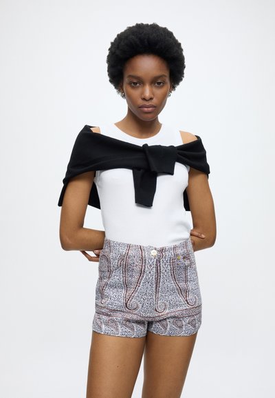 Femme aux cheveux courts et bouclés portant un haut blanc sans manches, un pull noir noué autour des épaules et un short à motifs, debout les mains dans le dos.