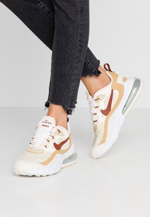 Stopy w beżowo-białych butach sportowych Nike Air Max z brązowymi akcentami i czarnych dżinsach z postrzępionym brzegiem na jednolitym, szarym tle.