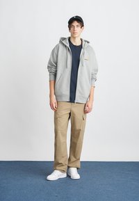Sweat à capuche gris zippé avec une poche avant, porté sur un t-shirt bleu marine, associé à un pantalon cargo beige et des baskets blanches. Casquette noire sur la tête.