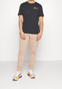Svart kortärmad t-shirt med en guld Nike-logotyp, kombinerad med ljusbeige tapered sweatpants och vita sneakers med orangea och svarta detaljer.