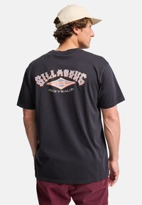 Billabong grafisk t-skjorte, svart bomull, kortermet, bakdesign med "AUSTRALIA" og logo, avslappet passform. Brukt med burgunder bukser.