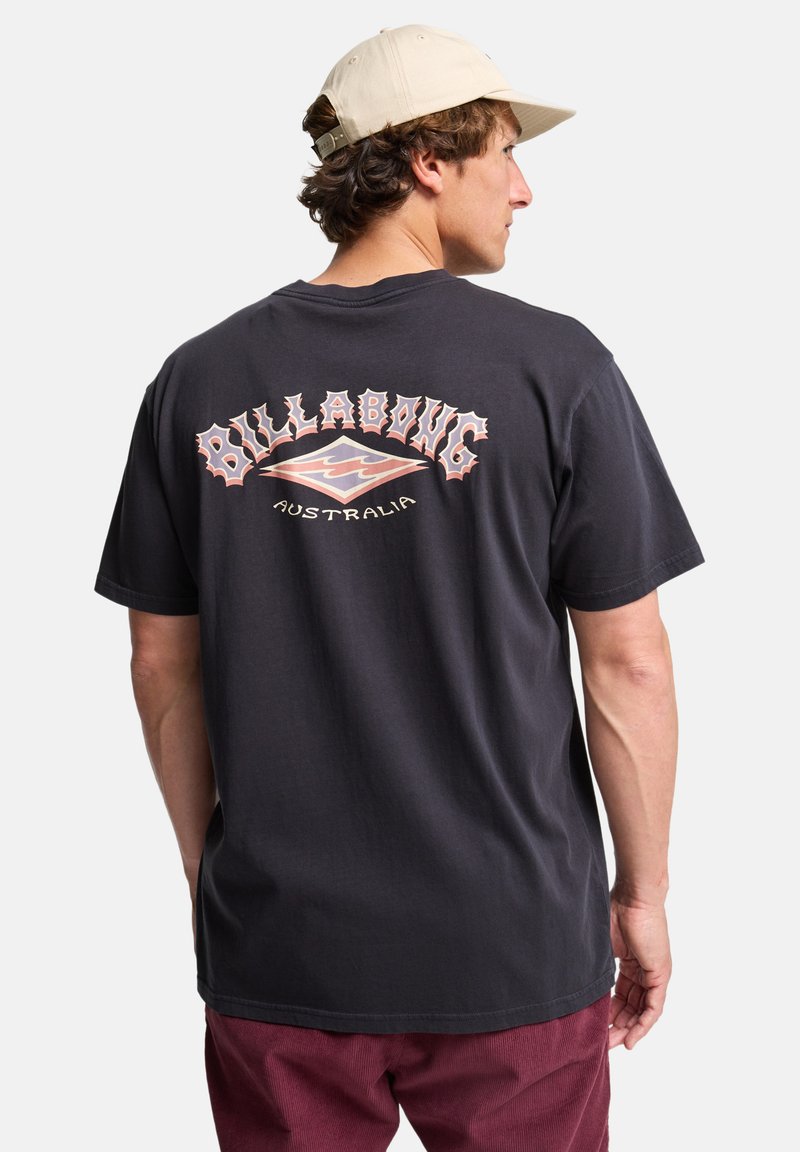 Billabong grafisk t-skjorte, svart bomull, kortermet, bakdesign med "AUSTRALIA" og logo, avslappet passform. Brukt med burgunder bukser.