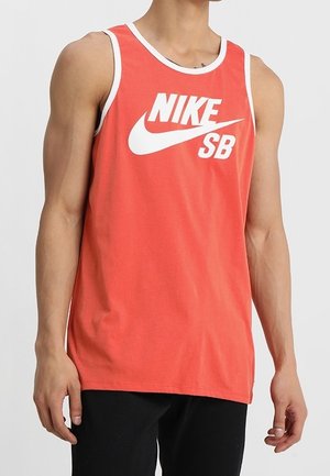 Mand iført rød ærmeløs tanktop med hvide kanter og hvidt "NIKE SB"-logo, kombineret med sorte bukser, stående mod en grå baggrund.