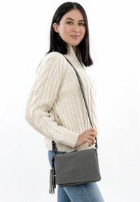 Grå läder crossbody-väska med texturerad yta, dragkedja högst upp och kvastdetalj. Modellen bär en krämfärgad kabelstickad tröja och jeanstight.