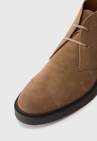 Botín de ante marrón con punta redonda, que cuenta con una suela plana de goma negra y un diseño de cordones minimalista para un aspecto limpio.