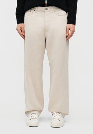 Άτομο που φοράει μπεζ παντελόνι φαρδύ στη μέση, λευκά αθλητικά παπούτσια και μαύρο τοπ με μακριά μανίκια, στέκεται μπροστά σε απλό φόντο.