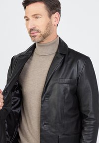 Czarna skórzana kurtka z satynowym wnętrzem, pojedynczą kieszenią na piersi, klapami i zapięciem na guziki, noszona na jasnobeżowym golfie.