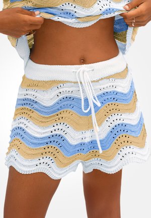 Persona che solleva un top beige, blu e bianco a maglia ondulata, indossando pantaloncini coordinati in maglia con vita con coulisse bianca.