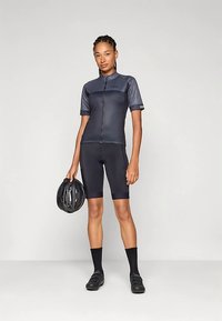 Κοντομάνικη φανέλα ποδηλασίας και σορτς σε σκούρο γκρι, κατασκευασμένα από ελαστικό ύφασμα. Κρατάει ένα μαύρο κράνος στο ένα χέρι, με μαύρα παπούτσια ποδηλασίας και κάλτσες.