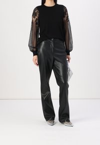 Pull noir avec manches bouffantes transparentes brodées, associé à un pantalon noir en similicuir taille haute et des chaussures argentées à bouts pointus.
