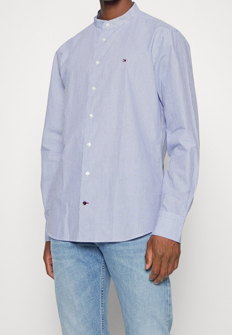 Homme portant une chemise légère à fines rayures bleu clair et blanc avec un col officier et un jean bleu clair.