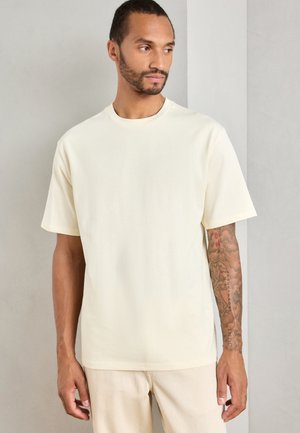 Man with tattooed right arm wearing a plain cream short-sleeve T-shirt and cream pants, looking to his left against a plain wall.