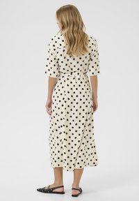 Abito lungo crema con pois neri, maniche corte e vita rigonfiata, abbinato a sandali neri flat. Tessuto dalla superficie liscia.