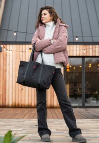 Padded pink jacket, white knit sweater, black tote bag with pink logo, black jeans, and gray boots. Background features wooden paneling and lights.