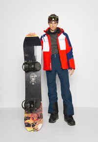 Snowboard mit schwarzem Oberteil, bunten Grafiken an der Unterseite, montierten schwarzen Bindungen. Modell in roter, blauer und grauer Jacke, blauen Hosen und schwarzen Stiefeln.