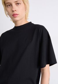 T-shirt in cotone nero con colletto alto e maniche corte. Presenta un marchio discreto sul petto e una vestibilità rilassata e ampia.