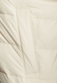 Beige pufferjacka med horisontell quiltad struktur. Har en rektangulär ficka integrerad i designen. Mjuk, slät yta.