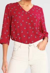 Femme portant un chemisier rouge à col en V avec un petit imprimé floral blanc et un jean beige taille haute, une main dans la poche.