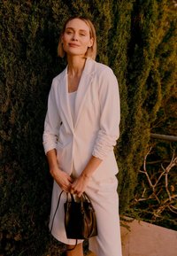 Femme en blazer blanc et jupe tenant un sac à main noir, debout à l'extérieur près d'un feuillage vert dense, souriant doucement.