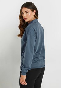 Felpa a maniche lunghe e colletto in misto cotone blu scuro, con polsini e orlo a costine. Modello mostrato da dietro, abbinato a leggings neri.