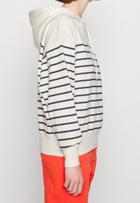 Beige hoodie met marineblauwe horizontale strepen. Voorzien van een trekkoord, ribgebreide manchetten en zoom. Gecombineerd met oranje broek.