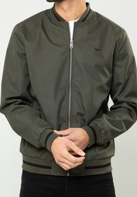 Homme portant une veste bomber verte olive zippée avec poignets et ourlet à rayures noires, les mains jointes devant, sur un fond uni.