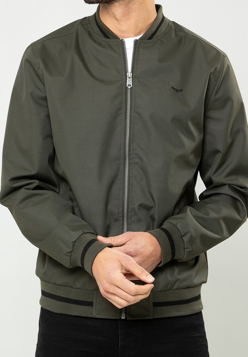Homme portant une veste bomber verte olive zippée avec poignets et ourlet à rayures noires, les mains jointes devant, sur un fond uni.