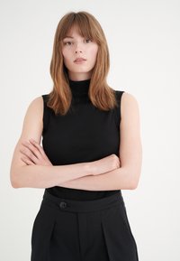 Haut à col montant sans manches noir côtelé associé à un pantalon noir taille haute avec un détail en bouton, sur un fond neutre.
