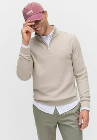 Beige stickad tröja med zip-krage, bärs över en vit randig skjorta, i kombination med gröna byxor. Vinröd keps med "SUNDAY"-logotyp.