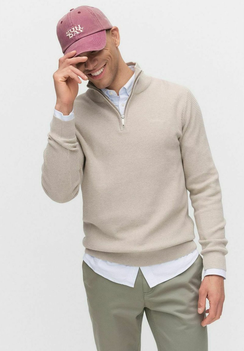 Beige stickad tröja med zip-krage, bärs över en vit randig skjorta, i kombination med gröna byxor. Vinröd keps med "SUNDAY"-logotyp.