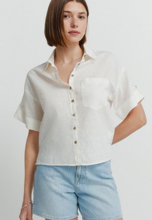 Junge Frau trägt ein weißes kurzärmeliges Hemd und hellblaue Jeansshorts, steht mit einer Hand in der Hüfte.