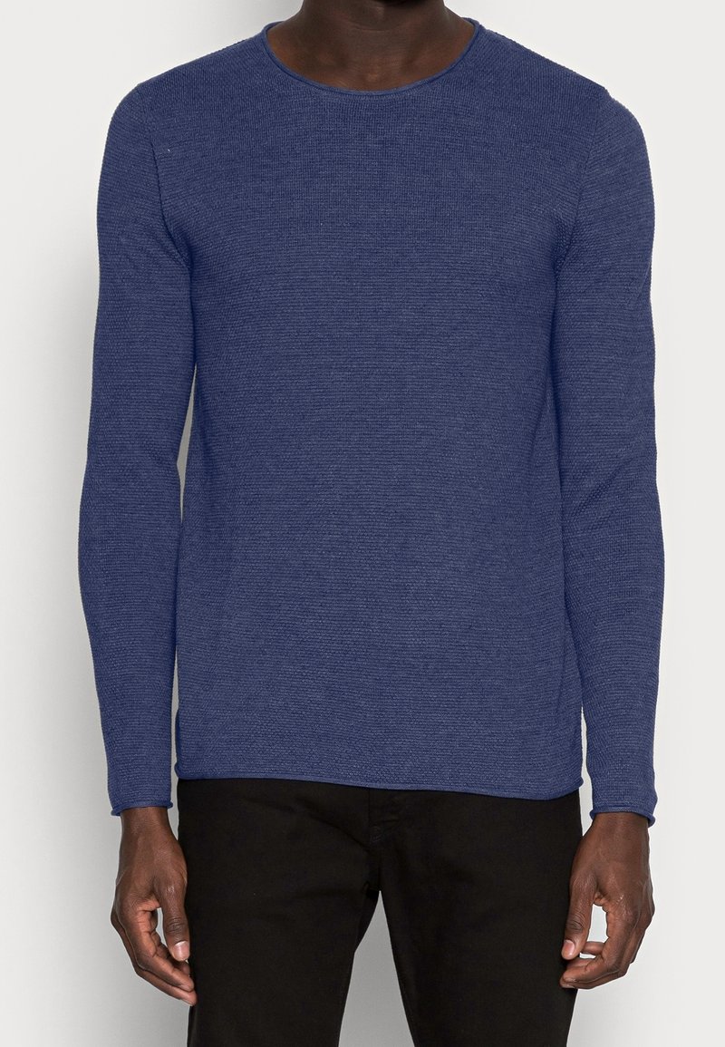 Pull à col rond à manches longues en tissu texturé bleu foncé, associé à un pantalon noir. Design simple, sans logos ni motifs visibles.