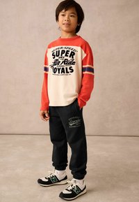 Langärmliges weißes und rotes T-Shirt mit grafischem Druck, kombiniert mit schwarzen Jogginghosen mit Logo. Die Schuhe sind schwarz-weiße Sneaker.