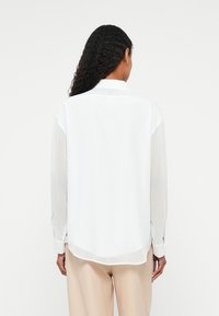 White long-sleeve shirt with a loose fit, made of lightweight fabric, featuring a back yoke detail and curved hemline.