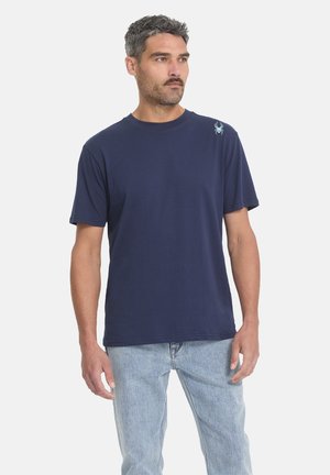 Man draagt een donkerblauw T-shirt met een klein wit spinnenlogo op de schouder en lichtblauwe spijkerbroek, staand tegen een effen achtergrond.