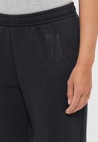 Pantaloni della tuta neri realizzati in tessuto morbido, con un elastico in vita e un sottile logo "GAP" in lettere scure sulla coscia sinistra.