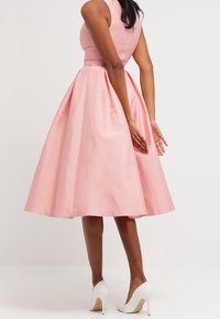 Vaaleanpunainen hihaton mekko, jossa on istuva yläosa ja levykkähelma, valmistettu sileästä kankaasta. Mekko ulottuu polveen ja sitä on yhdistetty valkoisiin korkokenkiin.