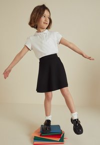 Weißes Polohemd mit Kragen und kurzen Ärmeln, kombiniert mit einem schwarzen, schwingenden Rock. Schwarze Lacklederschuhe und weiße Socken mit Rüschen.