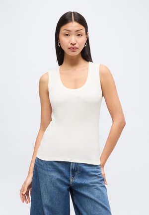 Młoda kobieta o długich ciemnych włosach nosi białą bluzkę bez rękawów i niebieskie dżinsy, stojąc na jasnym, jednolitym tle.