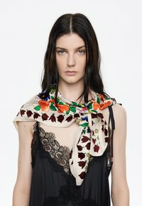 Bufanda de seda con patrones florales y de tulipanes coloridos sobre un fondo crema, drapeada alrededor del cuello, combinada con una blusa de encaje negra.