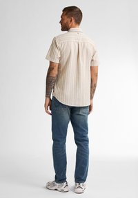 Camisa de botones de manga corta en beige con finas rayas verticales; combinada con jeans azules y zapatillas deportivas; corte relajado, dobladillo redondeado.