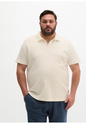 Mężczyzna z brodą, ubrany w kremową koszulkę polo z krótkim rękawem i granatowe spodnie, stoi z jedną ręką w kieszeni na białym tle.