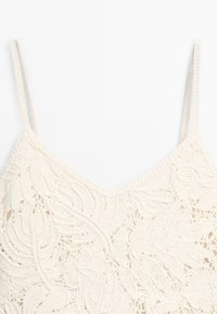 Weißes gehäkeltes Tanktop mit dünnen Trägern und filigranen Blumen- und Blättermustern auf hellem Hintergrund.