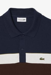 Granatowa koszulka polo z teksturowanej tkaniny, z białym i brązowym poziomym paskiem oraz zielonym logo krokodyla na piersi.