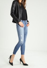 Mujer con chaqueta de cuero negra, camiseta negra, jeans ajustados rasgados azules y zapatos de tacón negro, de pie con las manos en los bolsillos.