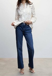 White lace blouse with a button front, paired with classic dark blue straight-leg jeans and white strappy sandals. Smooth textures and intricate patterns.