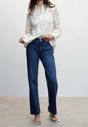 Blouse en dentelle blanche avec un devant à boutons, associée à un jean droit bleu foncé classique et des sandales blanches à lanières. Textures lisses et motifs complexes.