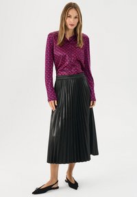 Camicia a maniche lunghe di un vivace rosa con un motivo geometrico, abbinata a una gonna plissettata in finta pelle nera e scarpe slingback nere.